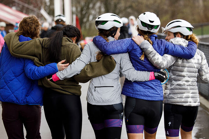 A group of female bike riders walk away with arms around each other.